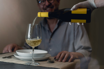 A sommelier pours white wine wearing protective latex gloves. A middle-aged adult returns to eat...