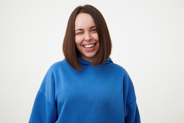 Portrait of young lovely brown haired woman without makeup giving cheerfully wink at camera and smiling broadly while standing over white background