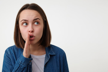 Fototapeta premium Portrait of young pretty blue-eyed brunette female covering her mouth with raised palm and looking carefully aside while telling secret story, posing over white background