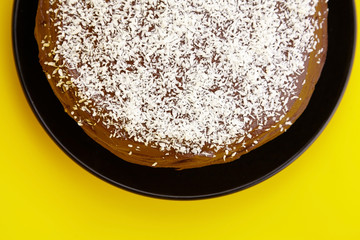 Chocolate cake decorated with flaked coconut, homemade pie on yellow background, top view. Home cake with cocoa ingredient on black plate