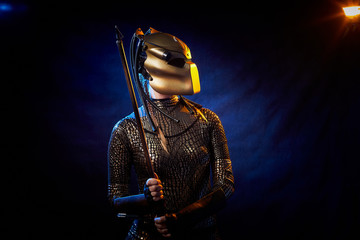 Valkyrie girl looking like an alien in shiny military armor, a strange helmet and a spear in a dark room with plants and vines. Model during a photo shoot, the actress during the shooting of the film.