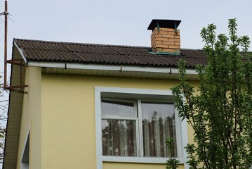 yellow attic of a private house with a white window under a gray slate roof and a brick chimney against the sky