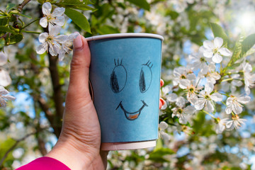 ecological cardboard mug with a happy smiley with hot coffee on the background of a blossoming cherry in the girlâ€™s hand