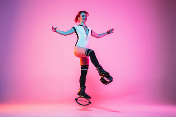 Fototapeta na wymiar Beautiful redhead woman in sportswear jumping in a kangoo jumps shoes isolated on purple-pink gradient studio background in neon light. Active movement, action, fitness and wellness. Fit female model.