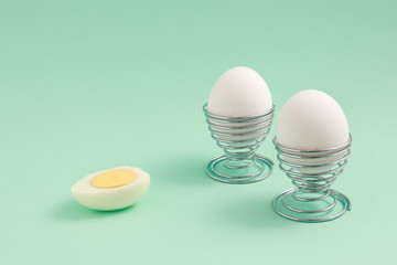 Two hard boiled eggs in metal egg cups and one half cut isolated on green background with copy space. 