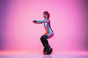 Beautiful redhead woman in sportswear jumping in a kangoo jumps shoes isolated on purple-pink gradient studio background in neon light. Active movement, action, fitness and wellness. Fit female model.