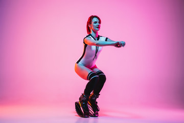 Beautiful redhead woman in sportswear jumping in a kangoo jumps shoes isolated on purple-pink gradient studio background in neon light. Active movement, action, fitness and wellness. Fit female model.