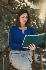 Obraz premium Smiling young woman sitting on a chair outdoor reading and holding a book. Open air activity concept. 