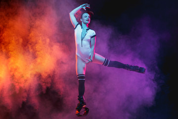 Beautiful redhead woman in sportswear jumping in a kangoo jumps shoes isolated on dark gradient studio background in neon lighted smoke. Active movement, action, fitness and wellness. Slender model.