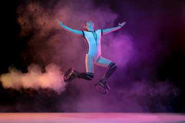 Fototapeta na wymiar Beautiful redhead woman in sportswear jumping in a kangoo jumps shoes isolated on dark gradient studio background in neon lighted smoke. Active movement, action, fitness and wellness. Slender model.