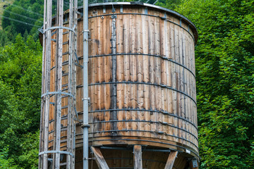 The wooden water supply tank on the background of green trees. The tank is placed on a wooden structure with pipes and a ladder attached to it. - Powered by Adobe