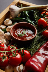 selective focus of basil leaves in tomato sauce near mushrooms, red cherry tomatoes, rosemary and chili peppers in wooden box