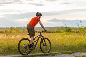 A cyclist in a bright orange T-shirt rides a bicycle along the meadow during sunset.