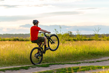A cyclist in a bright orange T-shirt rides on the rear wheel of a bicycle along a meadow during sunset.