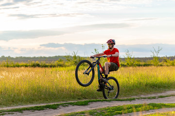 A cyclist in a bright orange T-shirt rides on the rear wheel of a bicycle along a meadow during sunset.