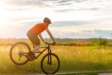 Fototapeta na wymiar A cyclist in a bright orange T-shirt stands on the front wheel of a bicycle along the meadow during sunset.