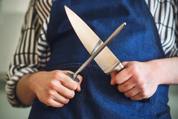 A cook in a blue apron and striped shirt is about to sharpen a kitchen knife on a long whetstone before cooking.