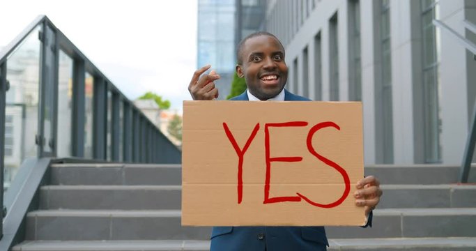 Portrait shot of African American male holding poster Yes and doing money gesture at demonstration or protest. Single protest outdoor. Activism concept. Cheerful man showing table with word yes.