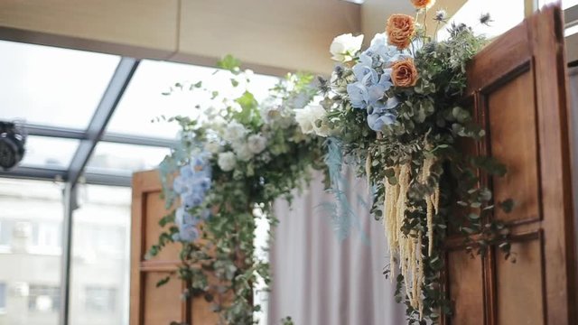Wedding altar with flower garland with silk curtains of multi-colored roses adorns door lintel at entrance to banquet hall for wedding. Oak door halves in the corridor with glass roof in the morning.