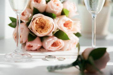 Wedding rings lie next to two glasses of champagne and a bouquet of roses in the sun