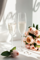 Wedding rings lie next to two glasses of champagne and a bouquet of roses in the sun
