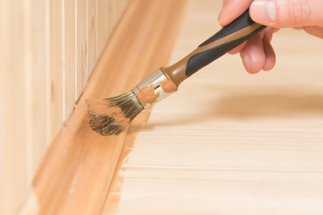 a small brush covers the skirting board of the wooden floor with dark varnish