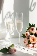 Wedding rings lie next to two glasses of champagne and a bouquet of roses in the sun