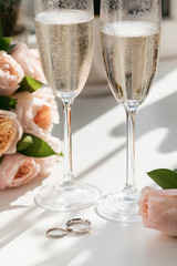 Wedding rings lie next to two glasses of champagne and a bouquet of roses in the sun