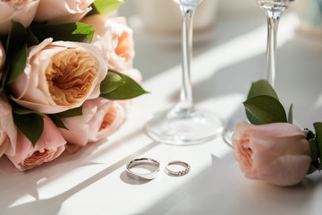 Wedding rings lie next to two glasses of champagne and a bouquet of roses in the sun