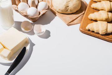 fresh croissants on wooden cutting board near raw dough, milk, butter and eggs on white background