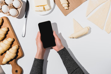 cropped view of woman holding smartphone with blank screen while cooking croissant on white background
