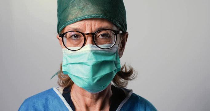Primo piano di un medico donna con occhiali, mascherina chirurgica e sfondo bianco.