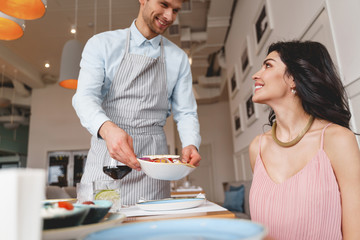 Joyful male waiter serving food to charming lady