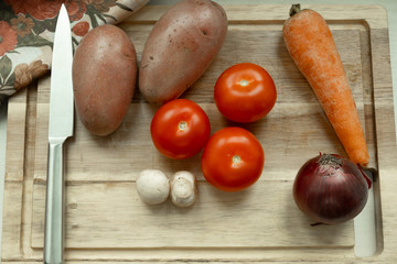  vegetables on a cutting board. Card on top of potatoes, tomato, carrots, red onions. mushrooms. The knife lies nearby.