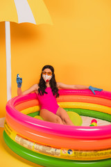 Obraz na płótnie Canvas young woman in bathing suit, sunglasses, latex gloves and medical mask sitting in inflatable pool and holding sanitizer on yellow