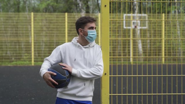 A male sportsman basketball player with a ball in a mask looks on the sides in a closed playground alone in quarantine during the pandemic