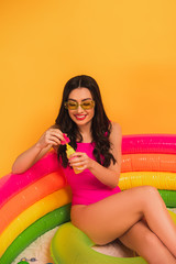 cheerful woman in swimsuit holding bottle with soap bubbles while sitting in inflatable pool on yellow