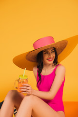 cheerful girl in straw hat and swimsuit holding glass with orange juice on yellow