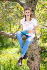 Smiling teenage girl in white T-shirt and jeans sitting on a tree, reading book on ebook reader. Great idea and activity to spend summer holidays in nature.