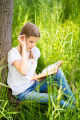 Teenage girl in white T-shirt sitting on the grass by the tree in the garden and reading interesting book. Great idea and activity to spend summer holidays in nature.