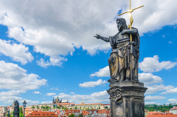 Fototapeta na wymiar Saint John the Baptist statue on Charles Bridge Karluv Most over Vltava river with Prague Castle, St. Vitus Cathedral in Hradcany district, blue sky white clouds background, Bohemia, Czech Republic