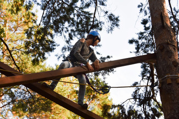 In a rope park, a beautiful young girl overcomes an obstacle at high altitude, climbs logs, amid huge trees and a clear sky, in a pine forest