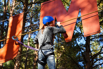 In a rope park, a beautiful young girl overcomes a difficult obstacle at high altitude, stands on a rope, a shaky structure, a little scary and exciting, against the background of huge trees and clear