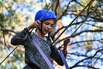 In a rope park, a beautiful young girl overcomes an obstacle at high altitude, a closeup portrait, excitingly, against the backdrop of huge trees and clear sky, in a pine forest