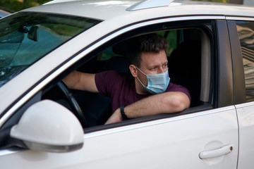 A man driving a car in a medical face mask during coronavirus outbreak, a taxi driver in a mask, protection from the virus. Driver in white car. Quarantine, covid-19.