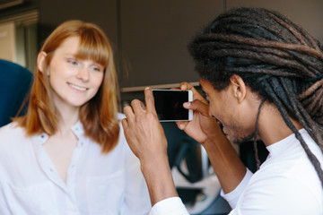 Ethnic African young man with dreadlocks taking a photo with a mobile phone of a redheaded girl in a kitchen