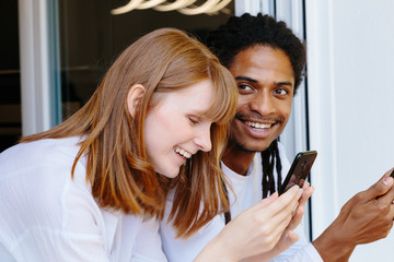 Two young smiling people of different ethnicities leaning on a window using the mobile phone