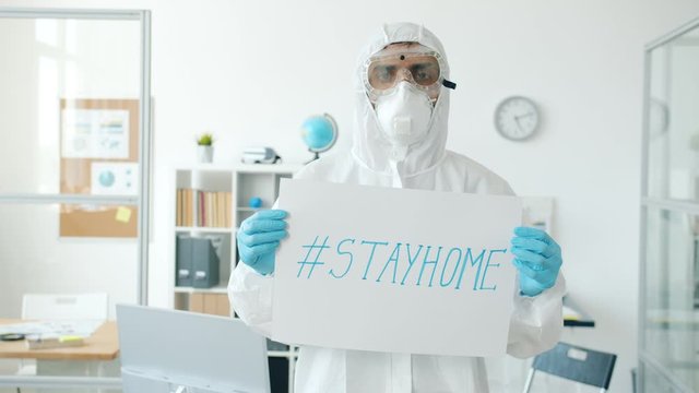 Portrait of Middle Eastern man in quarantine suit standing in empty office with stayhome banner and looking at camera. Covid-19, business and health concept.
