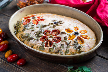 Homemade, beautiful and delicious, italian focaccia in round baking bowl. Decoration that looks like flowers in garden is made of cherry tomatoes, olives and fresh herbs.