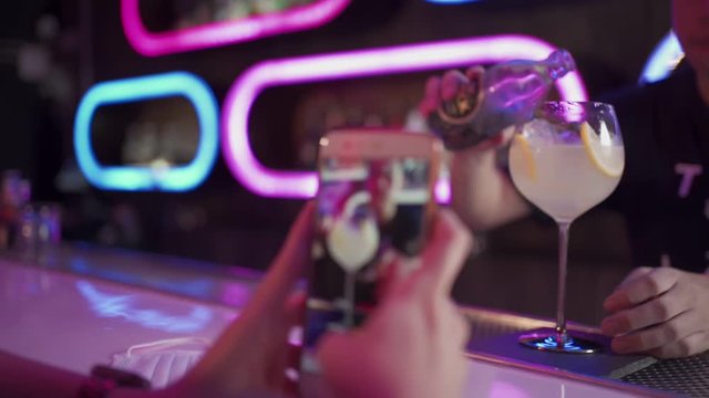 woman customer using smartphone taking photo of cocktail at nightclub bar, bartender mixing tasty drink, posting on social media timeline, female sipping on cocktail, bar counter, slow motion